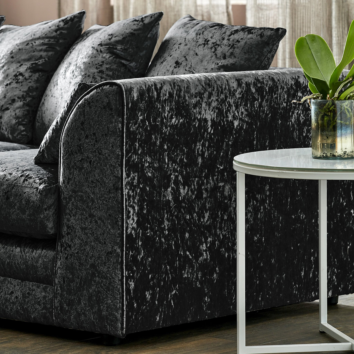 A crushed velvet sofa with plush pillows near a marble-topped side table with a plant, set against a curtained window.