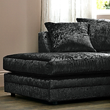 A plush, dark-colored chaise lounge with pillows rests in a room with beige walls and sheer curtains.