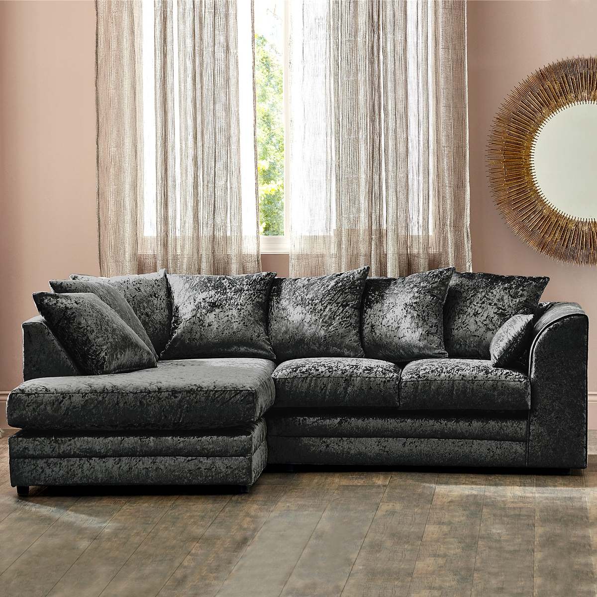 A plush, dark gray sectional sofa with multiple cushions; situated in a room with beige curtains, a window with a garden view, and a decorative golden mirror on the wall.