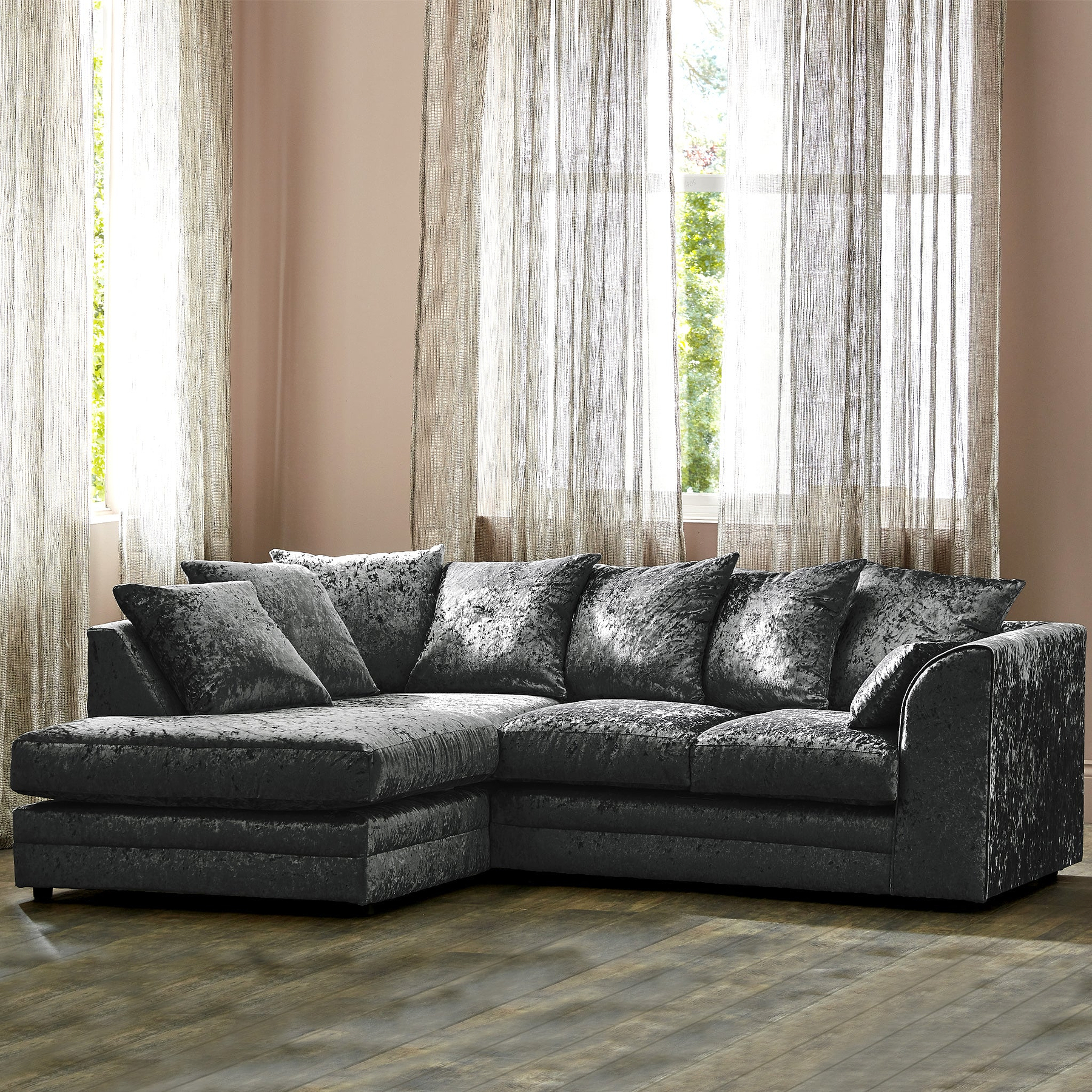 A crushed velvet corner sofa with cushions stands in a room with sheer curtains and a large window, creating a cozy interior setting.