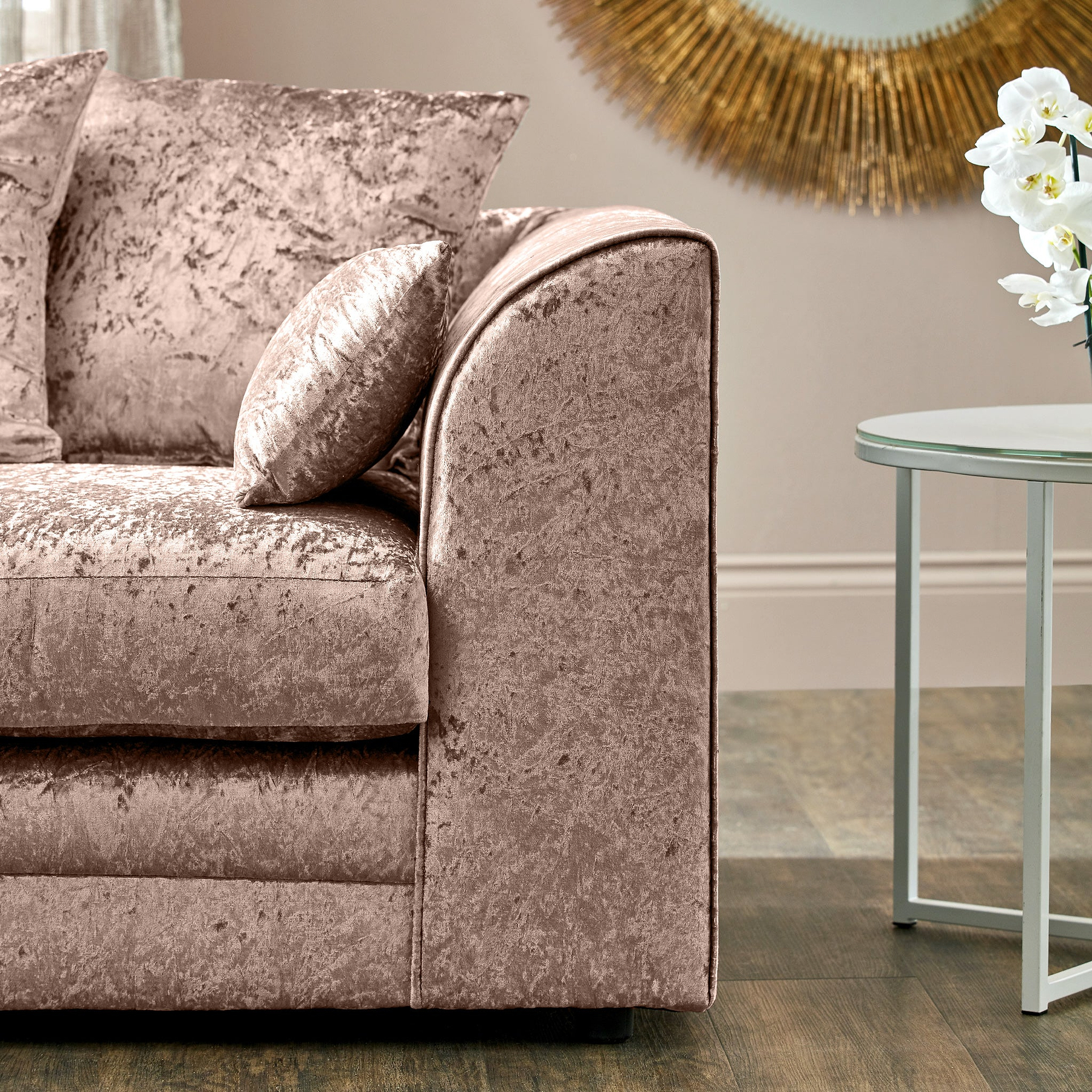 Velvet-textured, plush mauve sofa with matching pillows is resting, situated in a well-lit room with minimalistic decor, including a white side table and ornamental wall art.