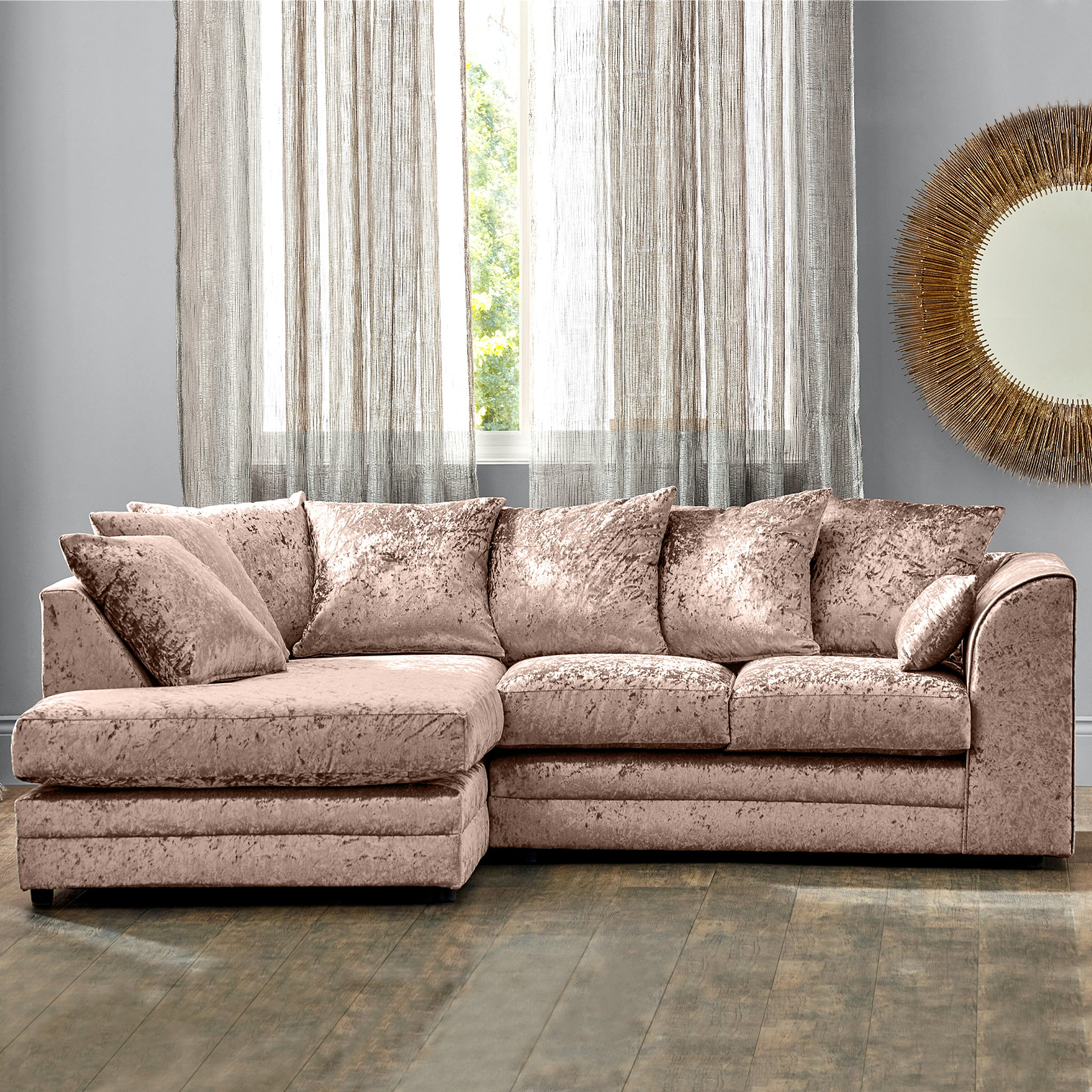 A plush L-shaped pink sectional sofa is positioned in a room with sheer curtains, wooden flooring, and a decorative mirror on the wall.