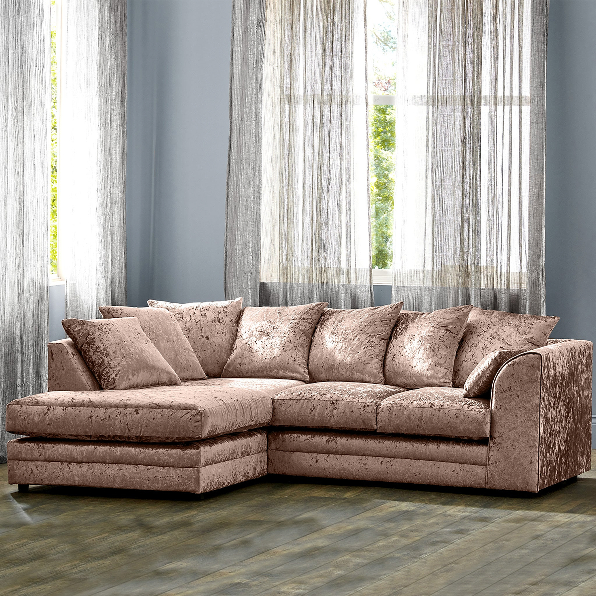 A plush pink sectional sofa sits in a room with sheer curtains, hardwood floor, and a blue wall.