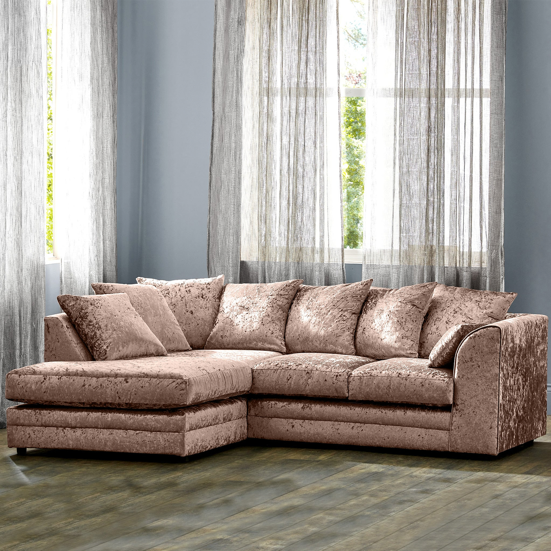 A plush pink sectional sofa sits in a room with sheer curtains, hardwood floor, and a blue wall.