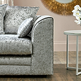 A plush silver velvet armchair with textured cushions, resting in a room with a beige wall and sunburst mirror. A white side table with orchids is nearby.