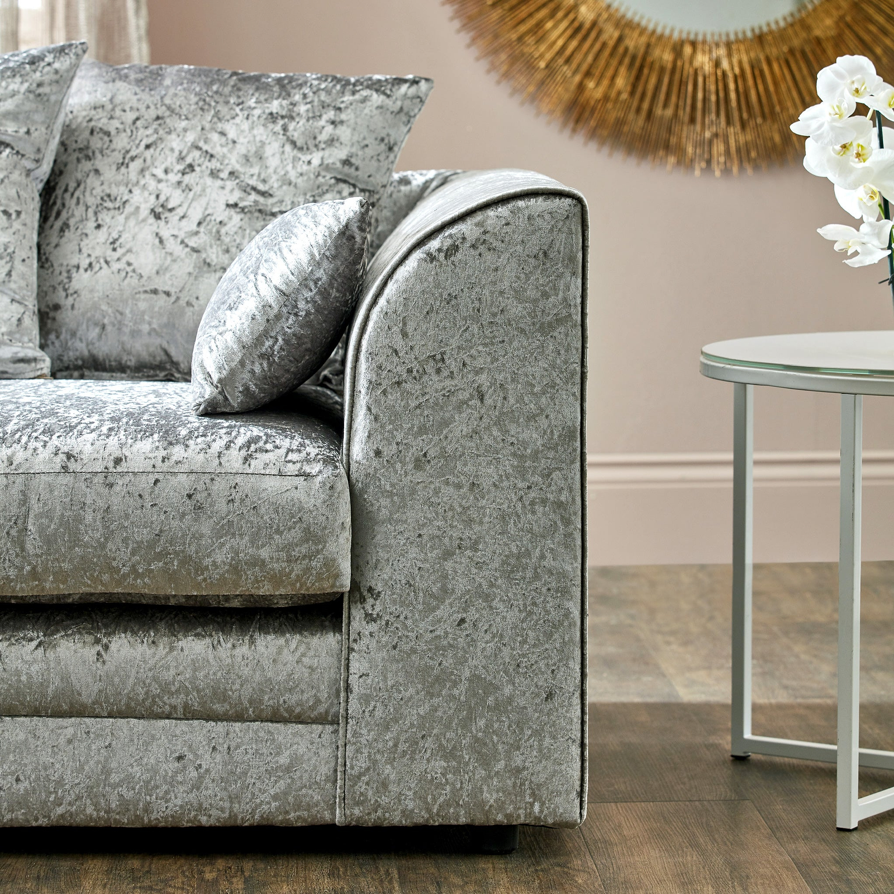 A plush silver velvet armchair with textured cushions, resting in a room with a beige wall and sunburst mirror. A white side table with orchids is nearby.