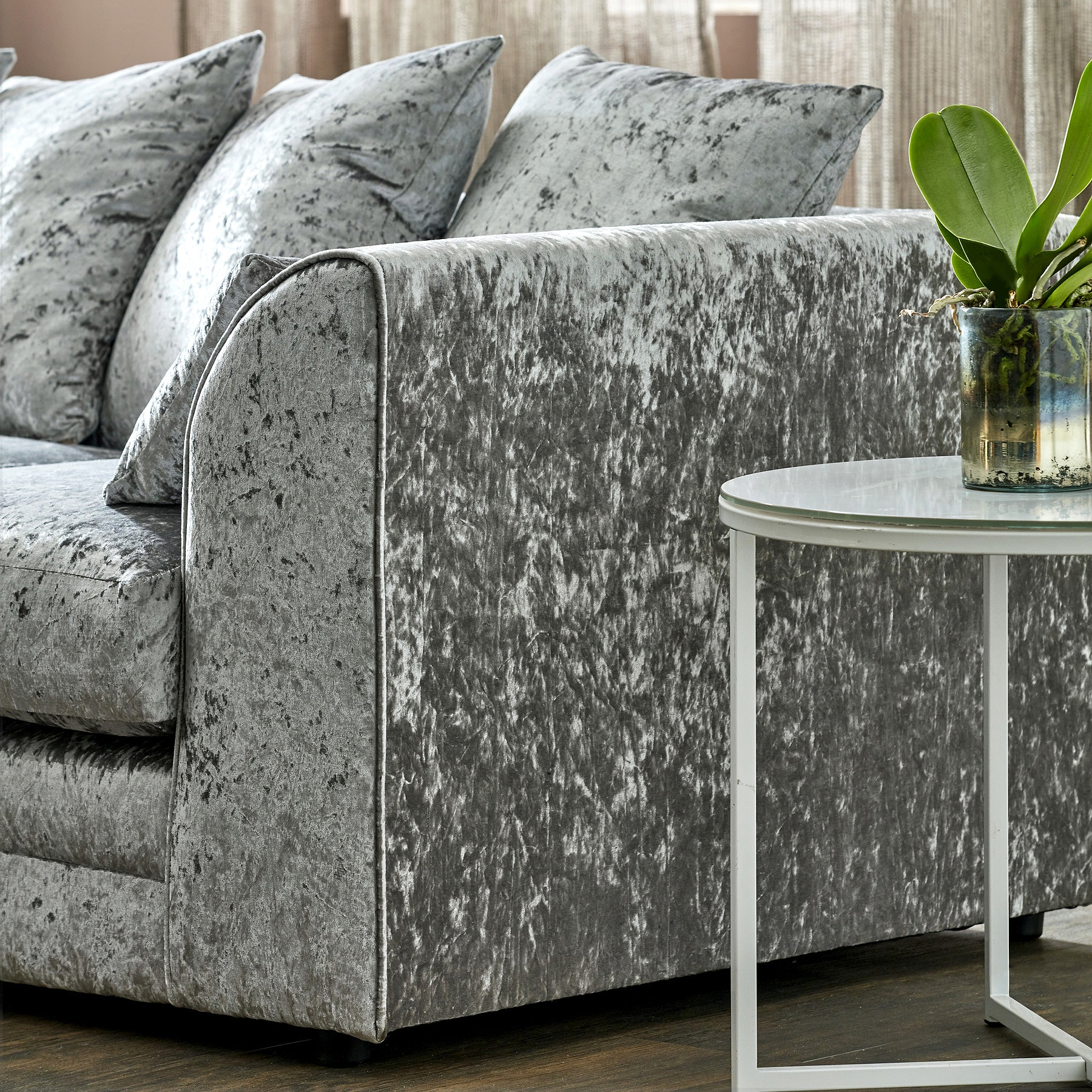 A velvet-textured, gray sofa with plush pillows sits idle, alongside a white marble-top table with a potted plant, arranged within a warmly lit, cozy living space.