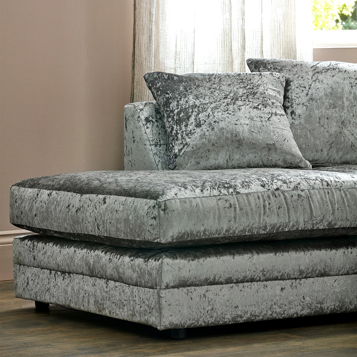 A grey crushed velvet loveseat with plush cushions is positioned in a well-lit room with sheer curtains.