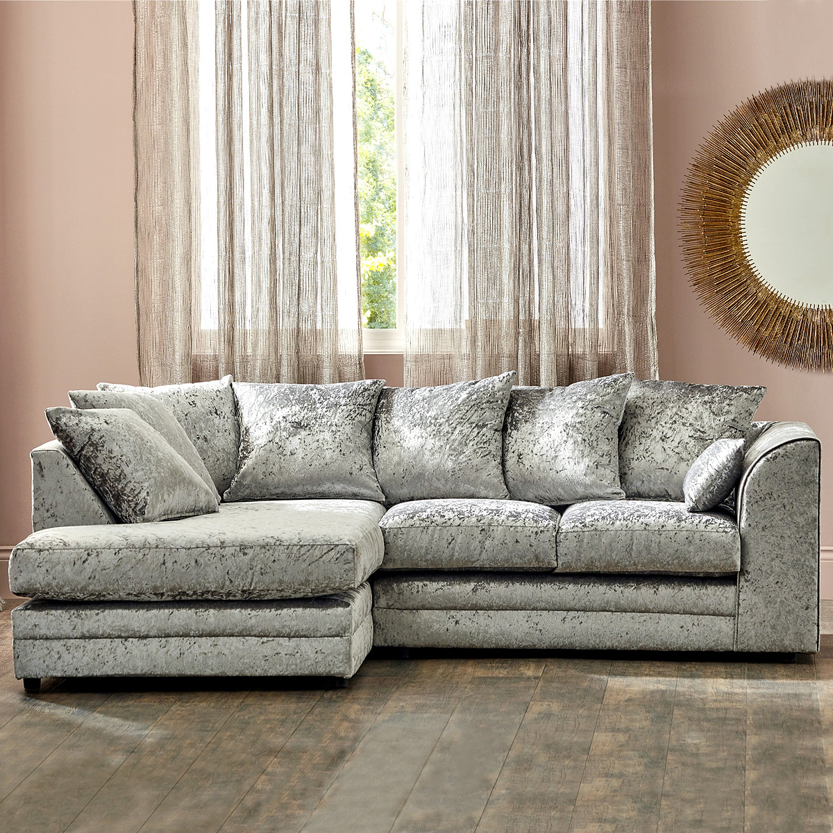 A plush L-shaped sectional sofa sits prominently, with patterned cushions, in a room featuring sheer curtains, a large round mirror, and hardwood flooring.