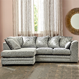 A plush L-shaped sectional sofa sits prominently, with patterned cushions, in a room featuring sheer curtains, a large round mirror, and hardwood flooring.