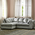 A plush, crushed velvet corner sofa with cushions stands in a warm-toned room with sheer curtains and a window behind it.