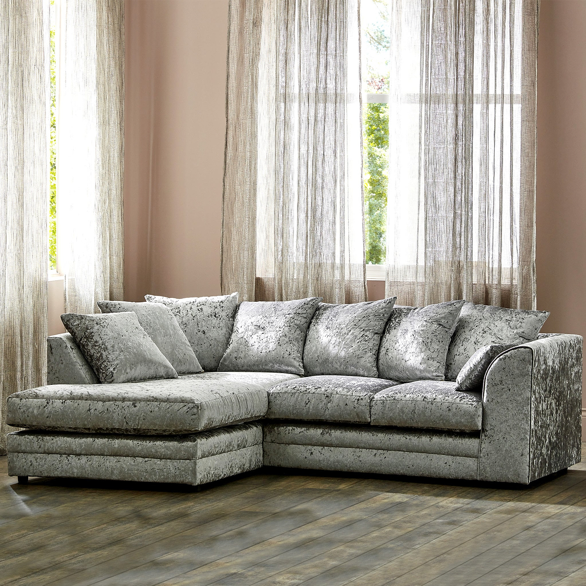 A plush, crushed velvet corner sofa with cushions stands in a warm-toned room with sheer curtains and a window behind it.