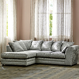 A plush, crushed velvet corner sofa with cushions stands in a warm-toned room with sheer curtains and a window behind it.