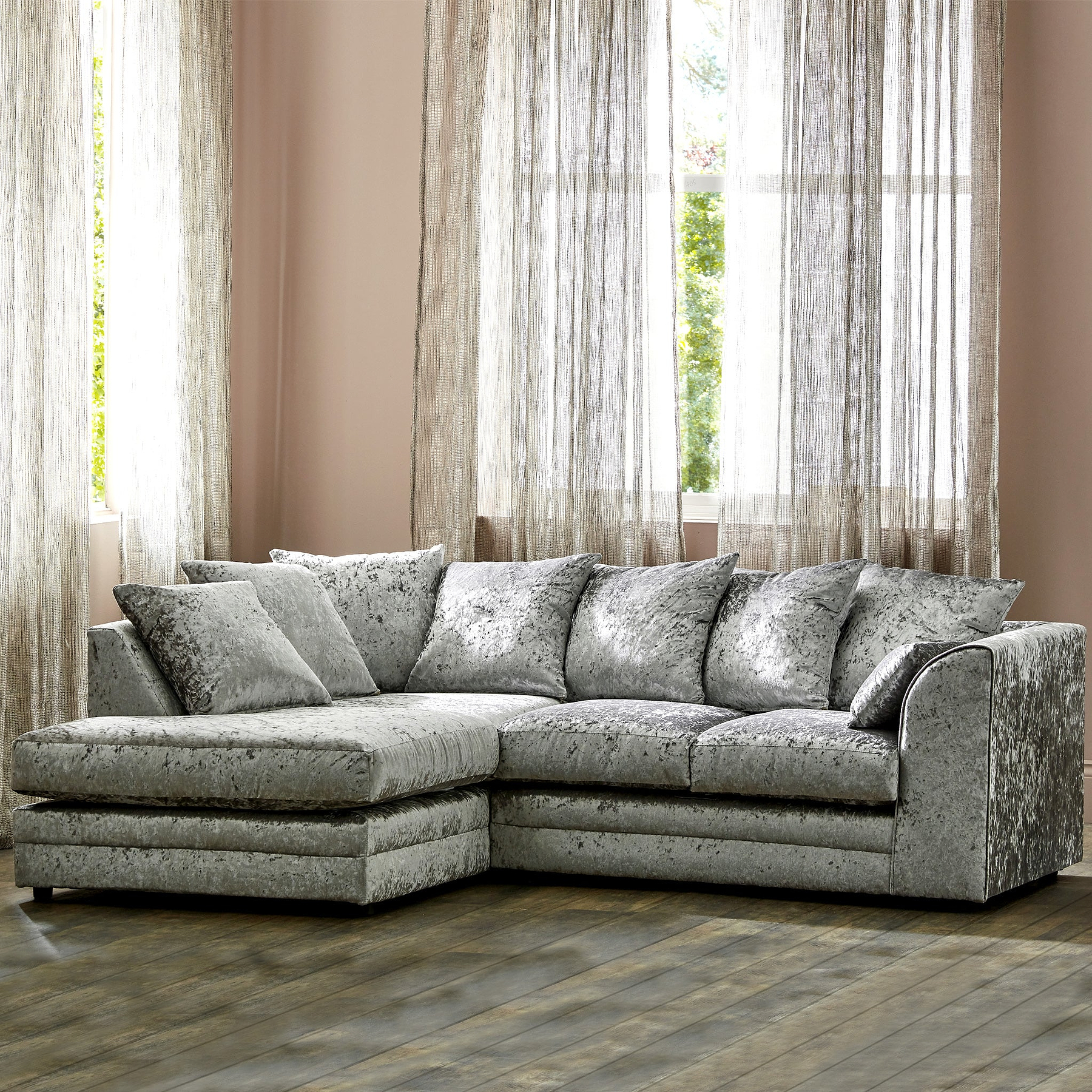 A plush, crushed velvet corner sofa with cushions stands in a warm-toned room with sheer curtains and a window behind it.