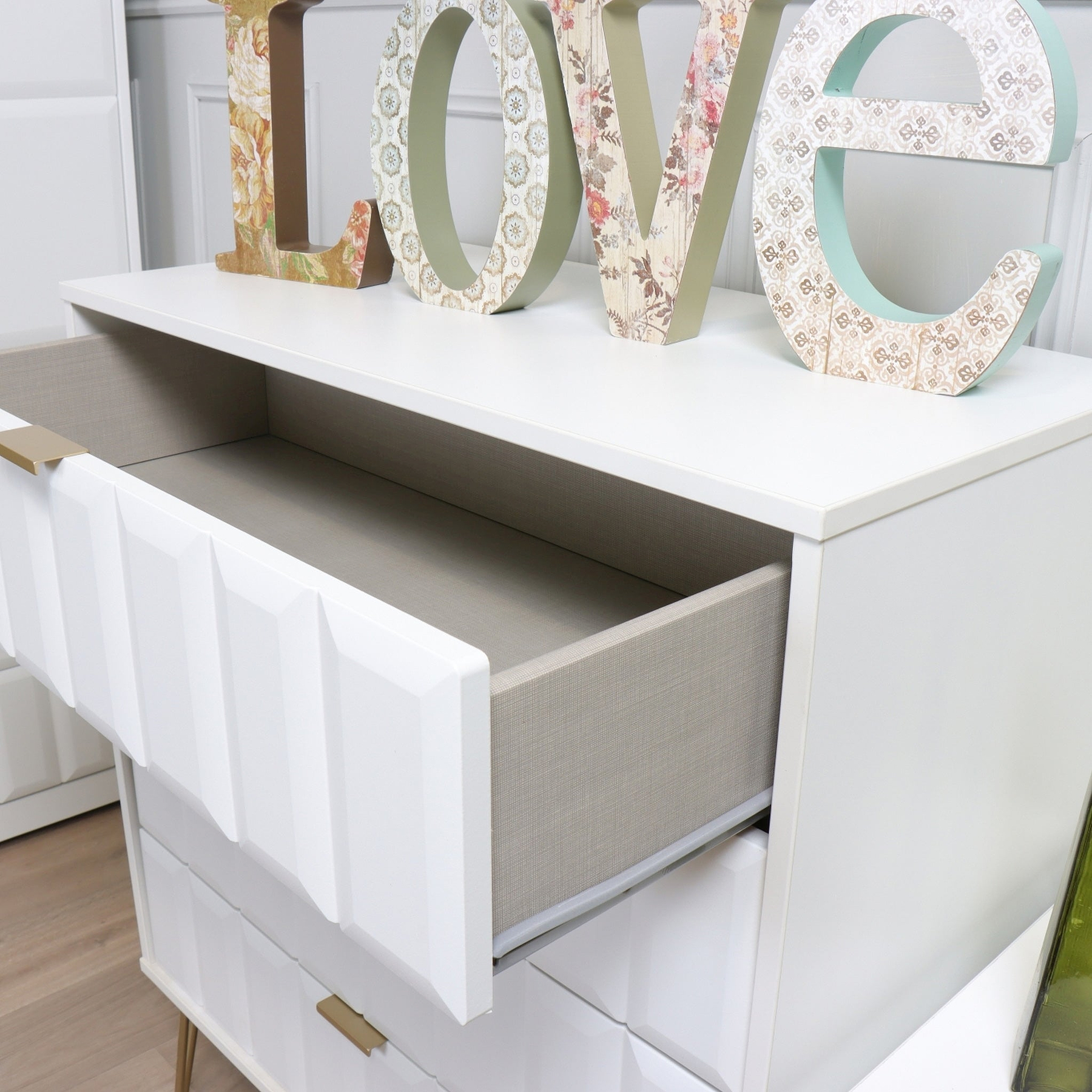Decorative letters spelling "LOVE" atop a white shelf with an open drawer revealing a fabric-lined interior against a soft-hued backdrop.