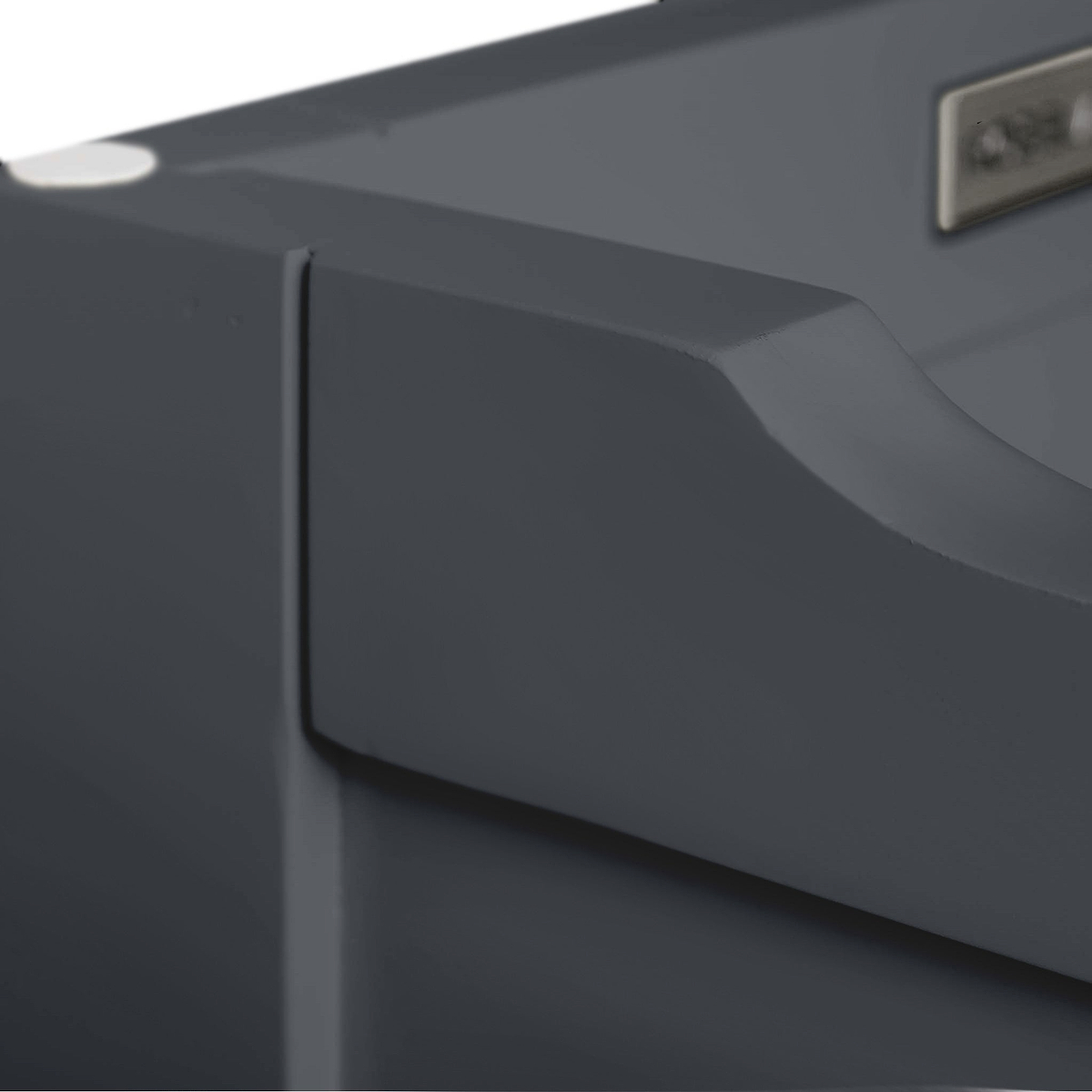 A close-up of a smooth dark gray surface with a curved edge and a metallic logo, possibly part of electronic equipment or furniture, against a blurred background.