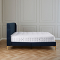 A single bed with a blue upholstered headboard and a white mattress, stands idle against a paneled wall in a room with herringbone wood flooring.