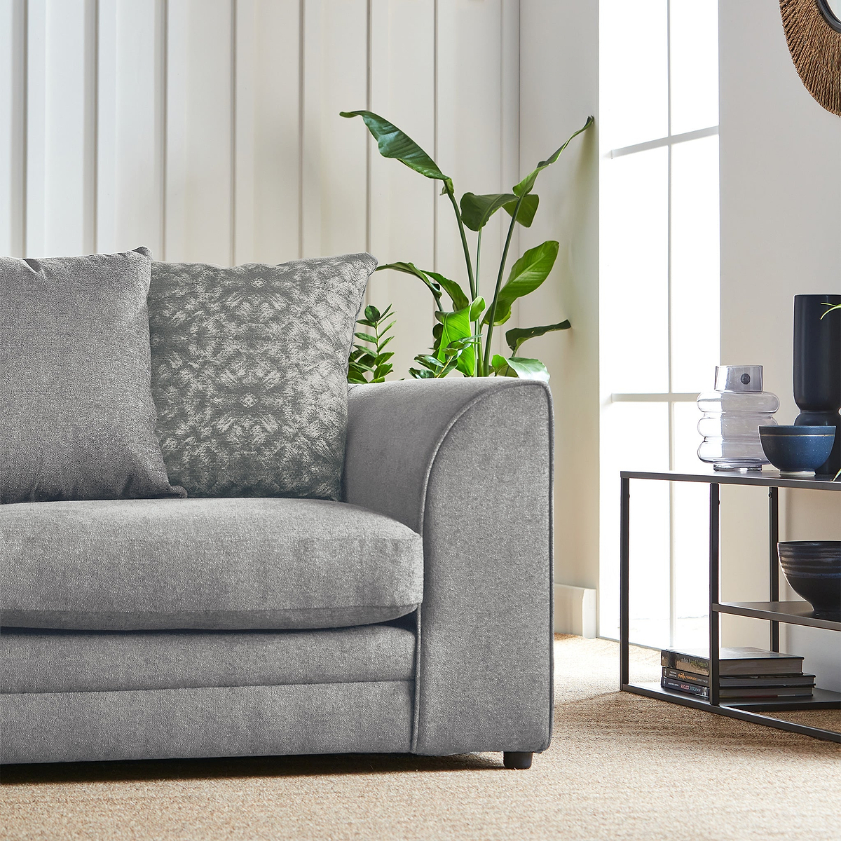 A grey sofa with patterned pillows sits beside a houseplant, with a side table holding decorative items near a window in a well-lit room.