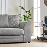 A grey sofa with patterned pillows sits beside a houseplant, with a side table holding decorative items near a window in a well-lit room.