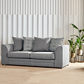A grey fabric sofa with cushions sits idle against a vertical striped wall, accompanied by a potted plant on the right, in a well-lit interior space.