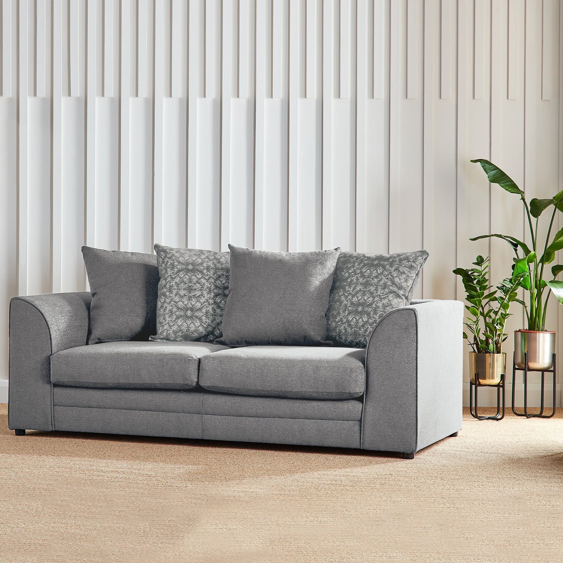 A grey fabric sofa with cushions sits idle against a vertical striped wall, accompanied by a potted plant on the right, in a well-lit interior space.