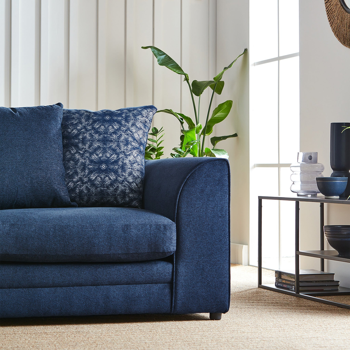 A navy blue couch with decorative pillows is situated beside a window with a green potted plant, near a side table with decorative items, in a well-lit, cozy room.