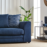 A navy blue couch with decorative pillows is situated beside a window with a green potted plant, near a side table with decorative items, in a well-lit, cozy room.