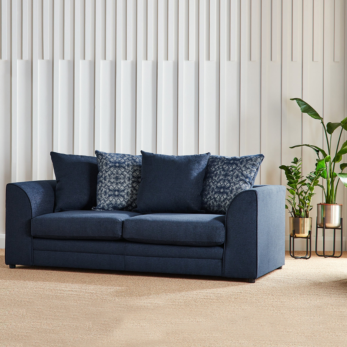 A dark blue sofa with patterned pillows rests against a white vertical slat wall, with a potted plant to the right.