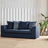 A dark blue sofa with patterned pillows rests against a white vertical slat wall, with a potted plant to the right.