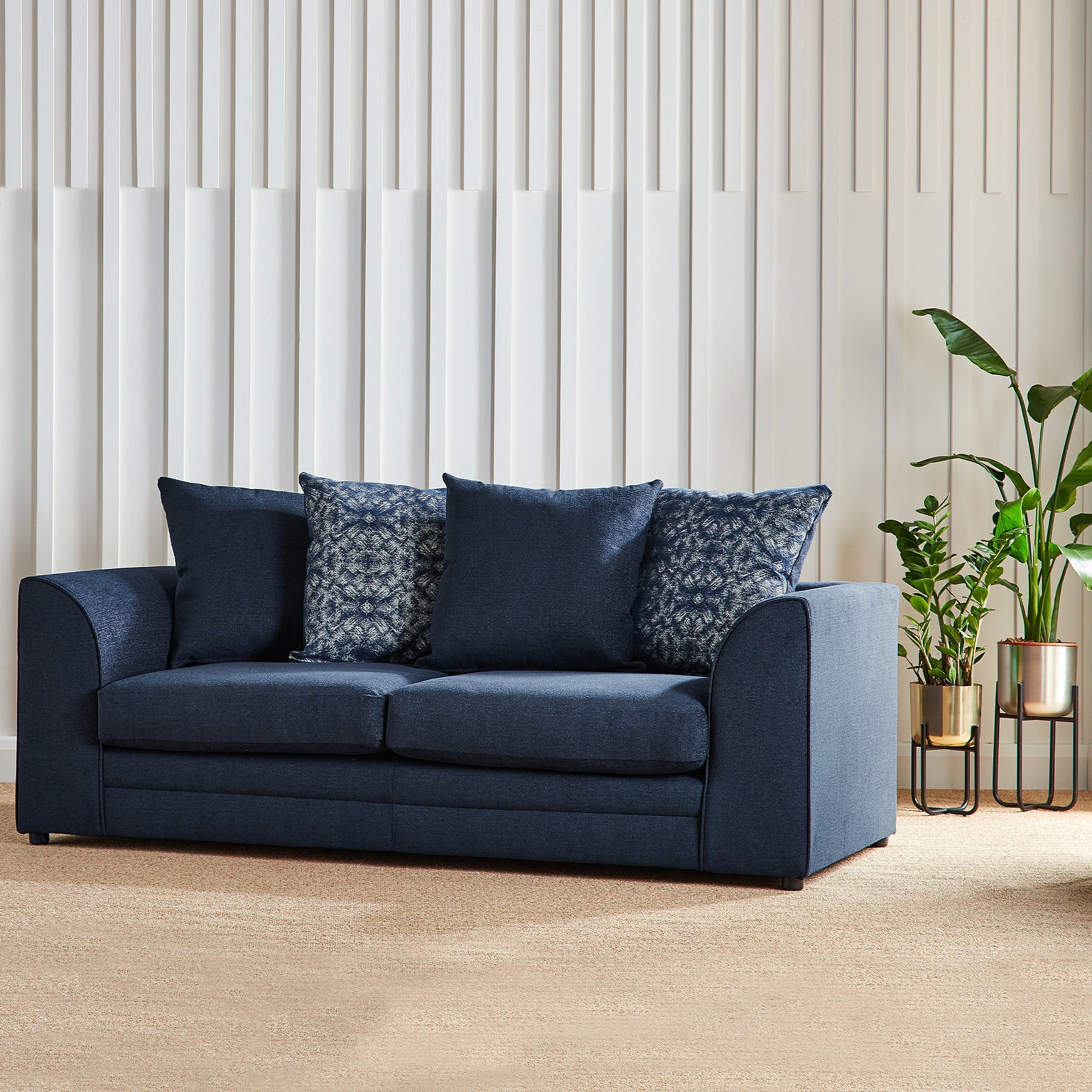 A dark blue sofa with patterned pillows rests against a white vertical slat wall, with a potted plant to the right.