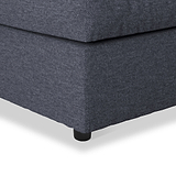 A gray upholstered ottoman with a cushion on top, featuring black rounded feet, sitting against an isolated white background.