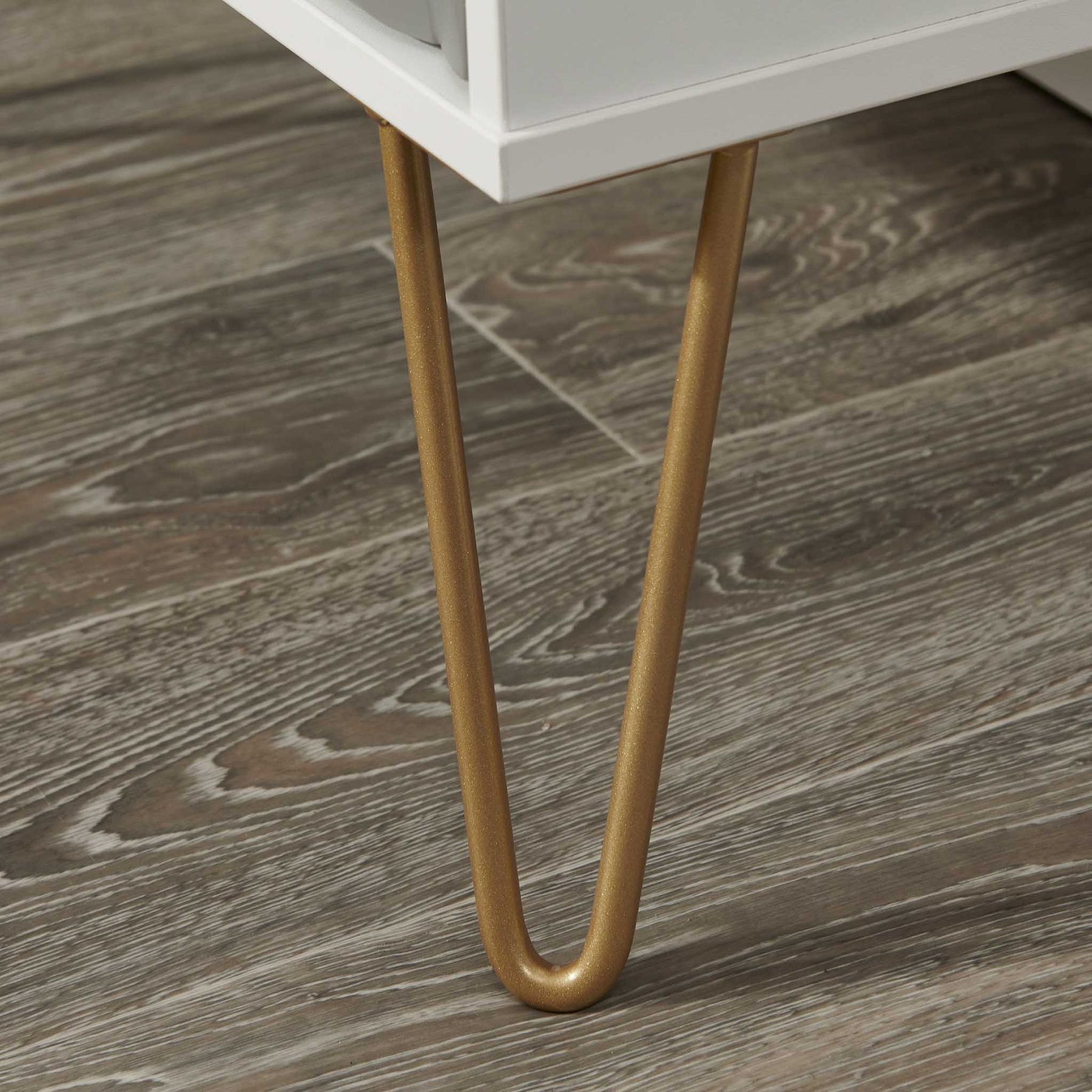 Golden-colored metal legs of a white piece of furniture, probably a table, standing on a wooden floor with a distinct grain pattern.