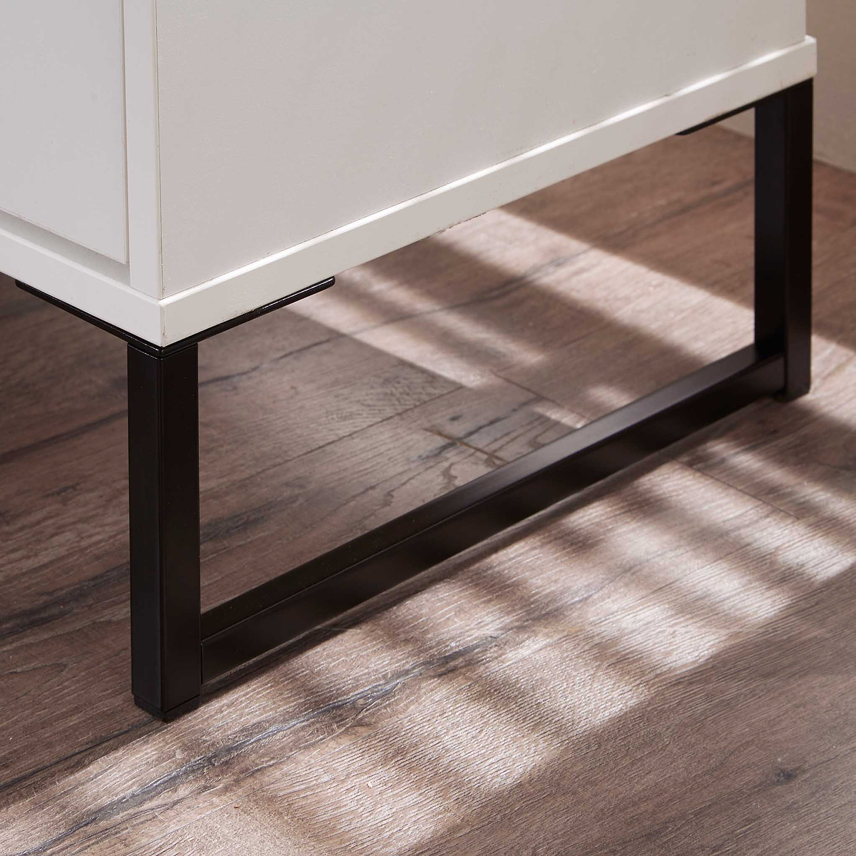 A white rectangular cabinet rests on black metal legs, casting soft shadows on a wooden floor with a striped light pattern from a window.