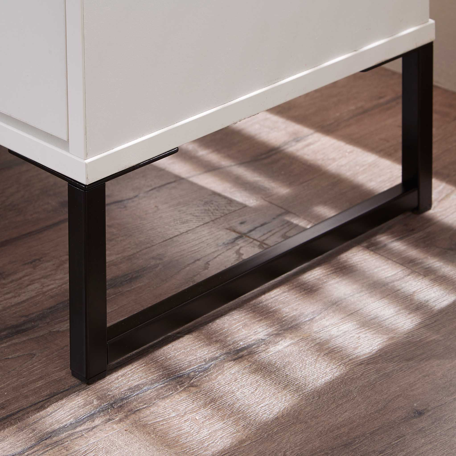 A white rectangular cabinet rests on black metal legs, casting soft shadows on a wooden floor with a striped light pattern from a window.