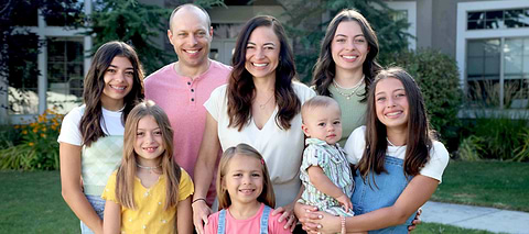 Smiling family of eight posing together outdoors in front of a suburban home.