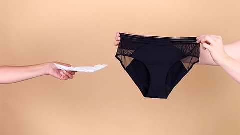 Hand offering a sanitary pad reaches toward black period underwear held against a beige studio background.