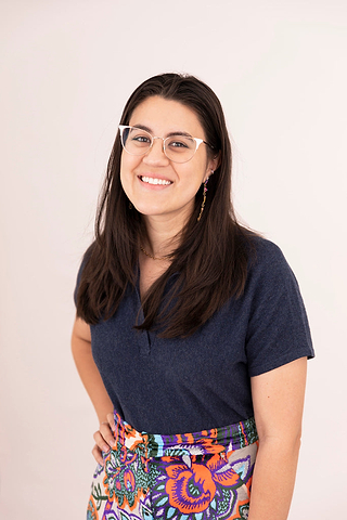 Smiling person with glasses wearing a navy shirt and colorful patterned skirt against a plain background.