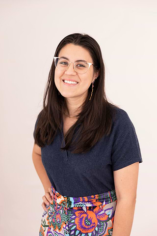 Smiling person with glasses wearing a navy shirt and colorful patterned skirt against a plain background.