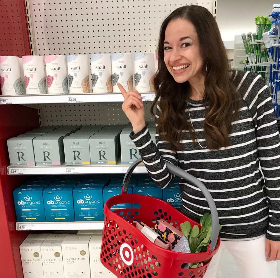 Smiling shopper pointing to menstrual cup products on a store shelf while holding a red basket.