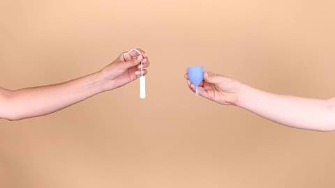 Hands holding a tampon and menstrual cup against a beige background.