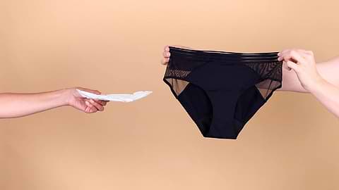 Hand holding a sanitary pad reaches toward black period underwear displayed against a beige background.
