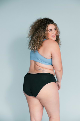 Curly-haired woman in a blue sports bra and black underwear smiles over her shoulder against a light blue background.