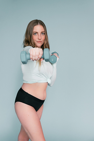 Woman in a white cropped top and black underwear holds dumbbells while posing against a light blue background.