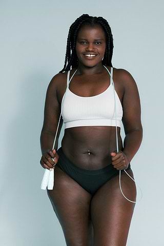 Smiling woman in a white sports bra and black underwear holds a jump rope against a light blue background.