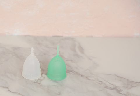 Two reusable menstrual cups, one translucent white and one green, displayed on a marble surface with a soft pink background.