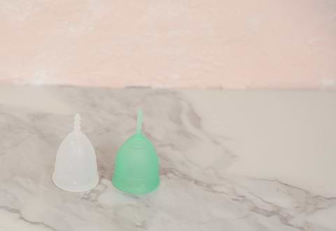 Two reusable menstrual cups, one translucent white and one green, displayed on a marble surface with a soft pink background.