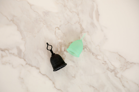 Black and green reusable menstrual cups placed on a light marble surface in a minimal product display.