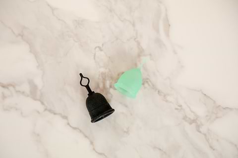 Black and green reusable menstrual cups placed on a light marble surface in a minimal product display.