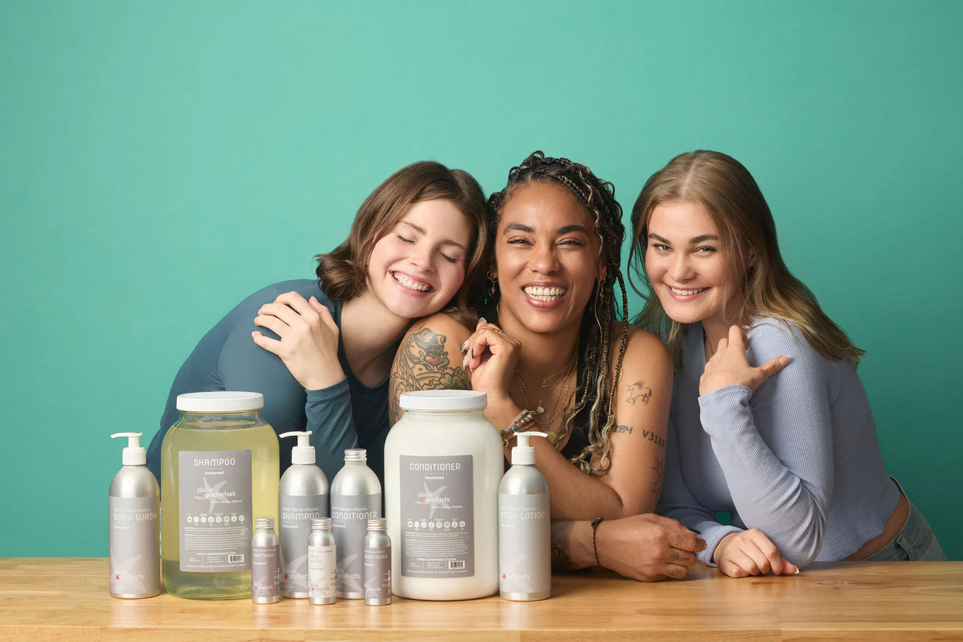 Three smiling women posing behind shampoo, conditioner, and body care products on a wooden table.