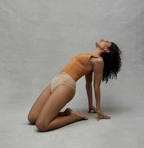 Woman in neutral-toned underwear and bodysuit kneels in a graceful pose against a gray studio backdrop.